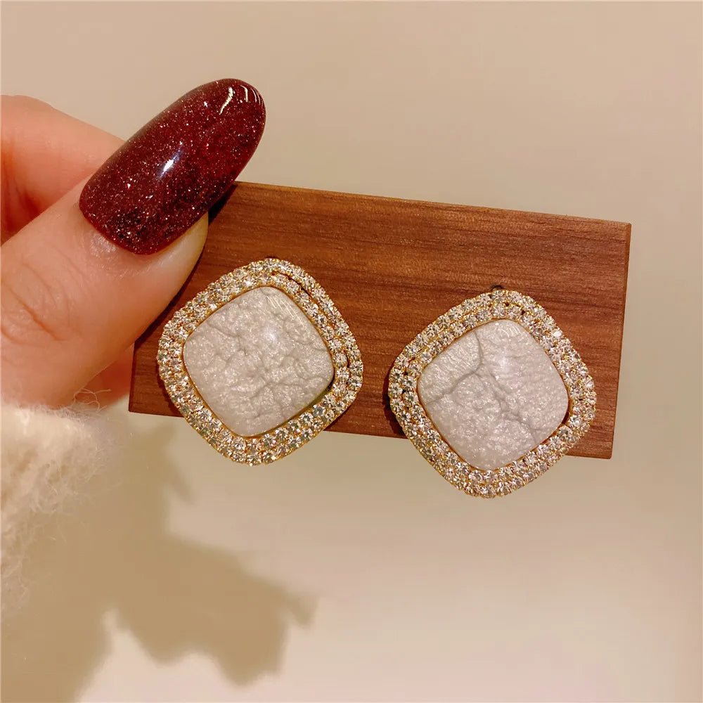 Square gold stud earrings with a white crackled stone center and pavé rhinestone border – elegant and modern statement jewelry